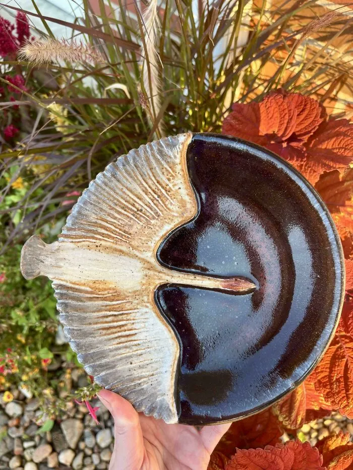 A person holding a unique pottery dish shaped like a mushroom with detailed gills and a dark brown glaze finish.
