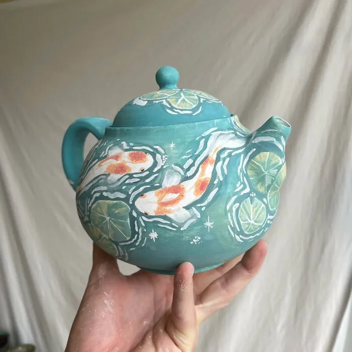 A hand holding a hand-painted light blue pottery teapot featuring orange koi fish and lily pads.
