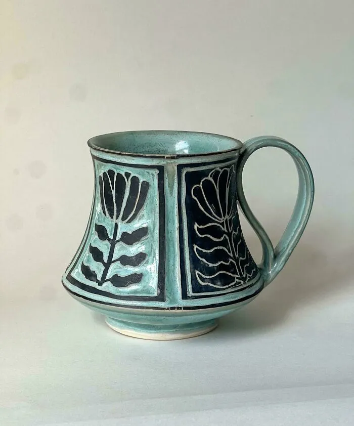 A light blue handmade pottery mug decorated with black floral patterns in relief.
