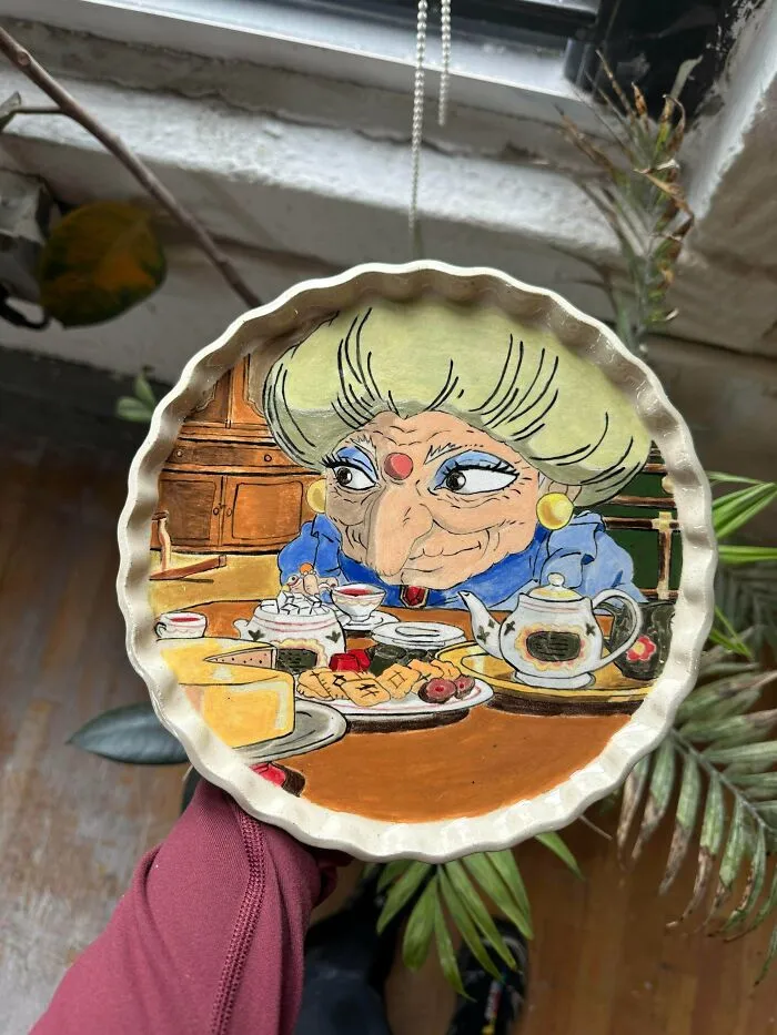 A hand holding a circular pottery dish featuring an intricate hand-painted scene of a witch at a tea table.