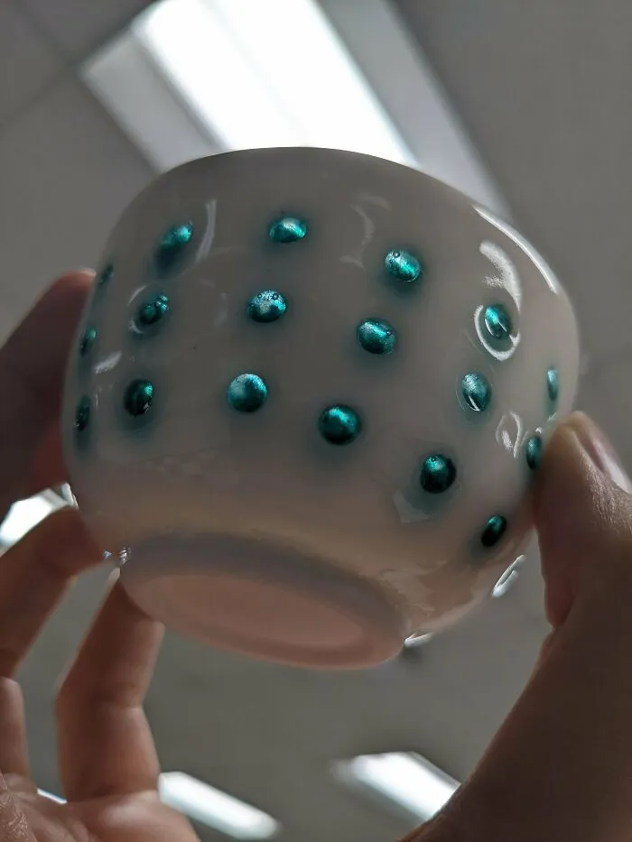 Hand holding a small white ceramic cup with raised teal dots, highlighting artistic pottery creations.