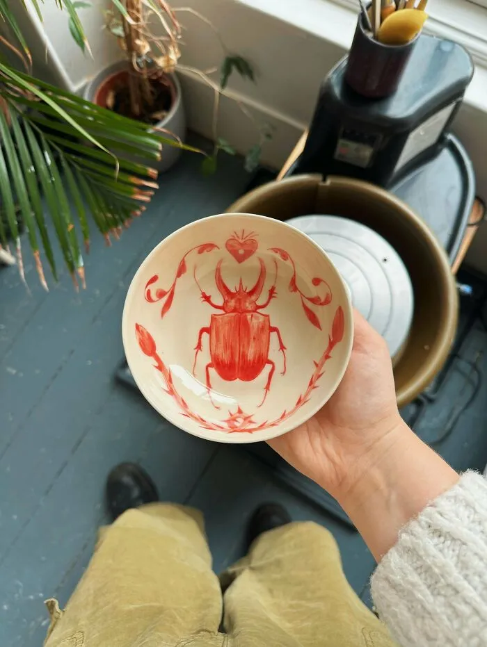 A hand holding a ceramic bowl featuring a detailed red beetle design, showcasing unique pottery creations.
