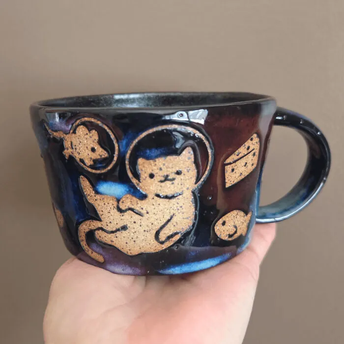 A dark glazed pottery mug decorated with a floating space cat and celestial details.
