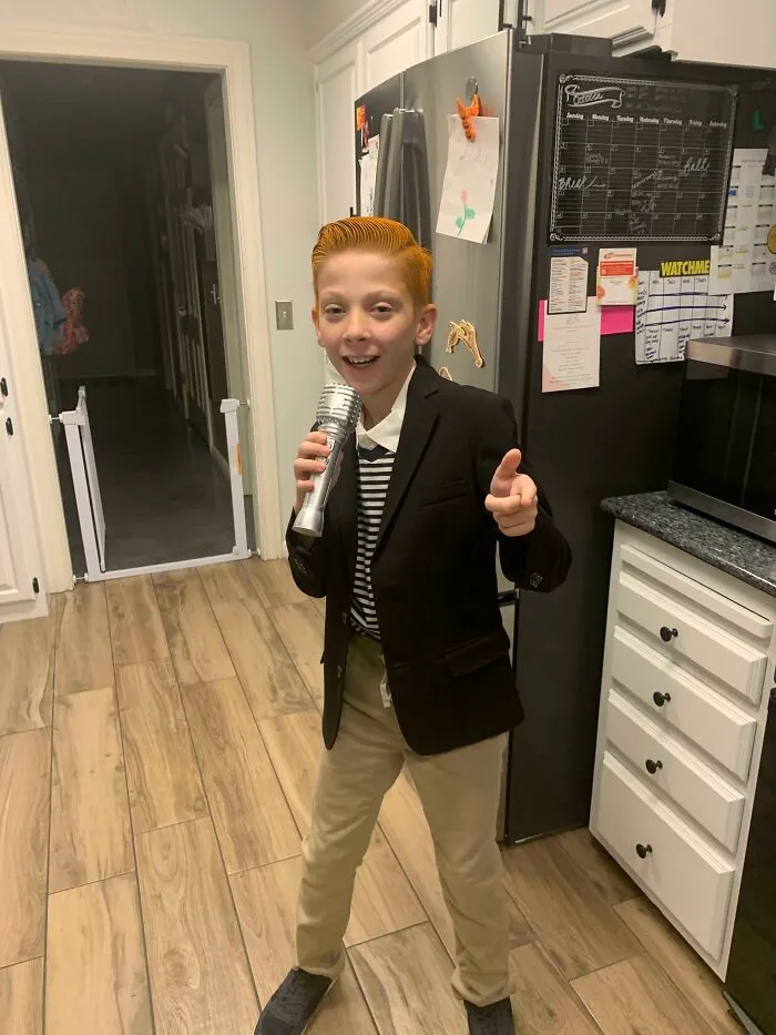 A young boy with styled orange hair dressed as Rick Astley holding a microphone for a hilarious kids Halloween costume.