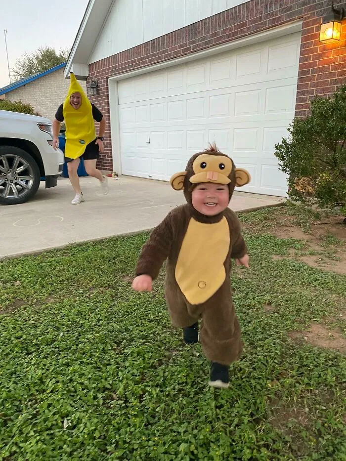 A toddler dressed in a monkey costume runs outside as an adult in a banana costume chases behind.