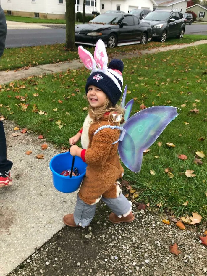 A happy toddler wearing a creative fairy bunny Halloween costume carrying a blue trick-or-treat bucket.