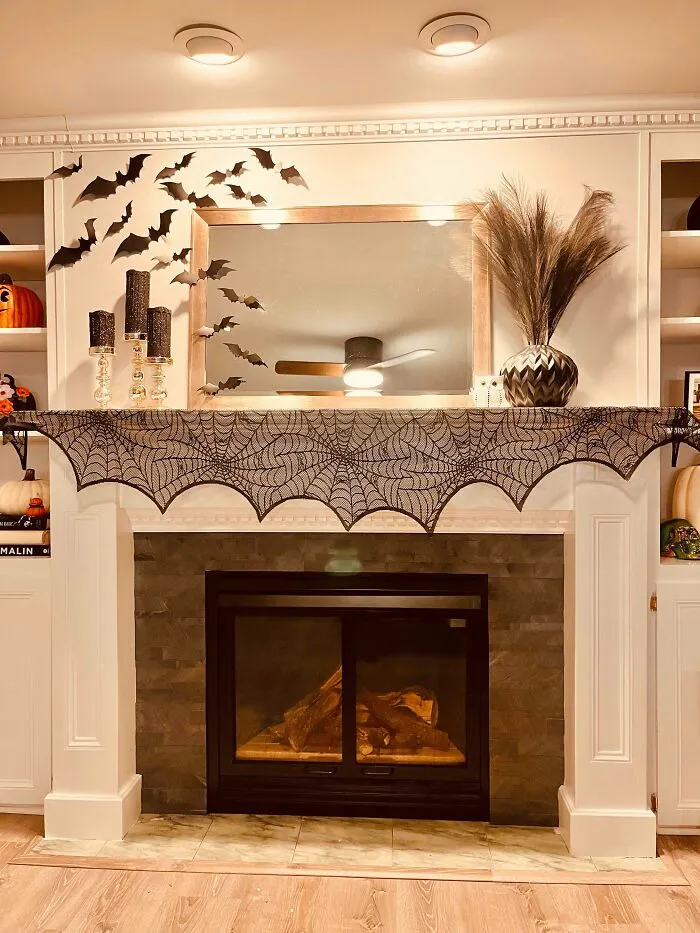 A fireplace mantel decorated for Halloween with a black spiderweb runner, wall bats, and dark candles