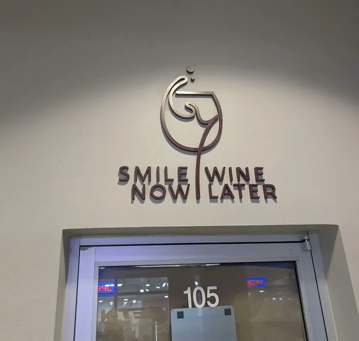 A terribly designed sign above a door that reads Smile Now Wine Later in a confusing layout