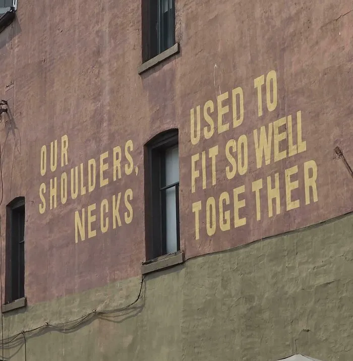 A terribly designed sign painted on a brick wall with confusingly layout text reading Our Shoulders Necks