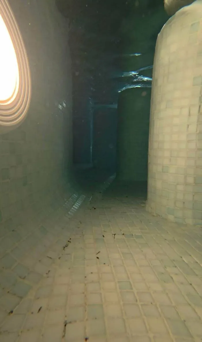 Eerie tiled hallway in an underwater pool facility showing submerged objects and deep water structures