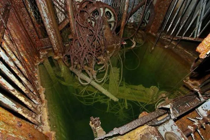 Dark underwater elevator shaft filled with murky green water and rusty cables, showcasing submerged objects.