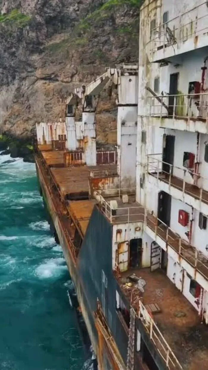 Massive rusty shipwreck tilted on its side near rocky cliffs, showcasing unnerving submerged objects.