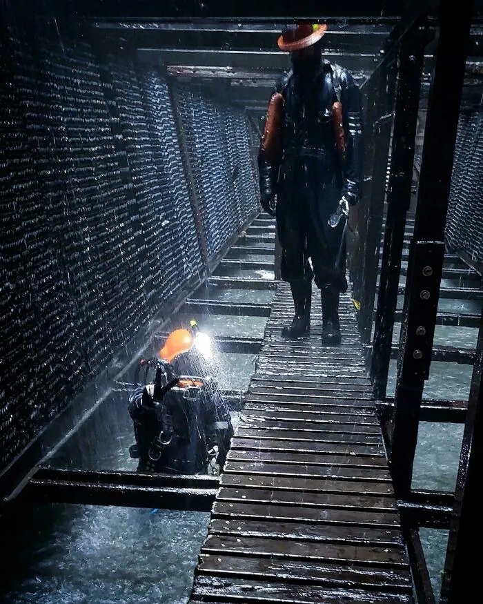 Workers navigating a narrow walkway over submerged objects in a dark channel