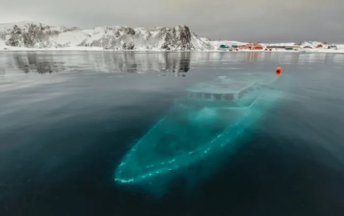 Eerie view of a yacht submerged objects visible under icy turquoise water near a snowy shore.