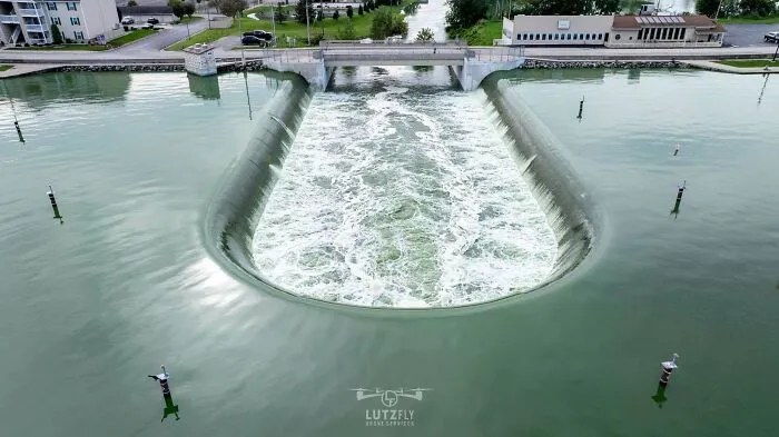 Aerial view of a U-shaped spillway creating an intimidating vortex in green water near a bridge.