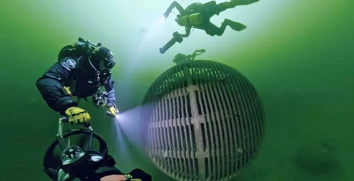Divers inspecting large submerged objects with flashlights under deep green water.