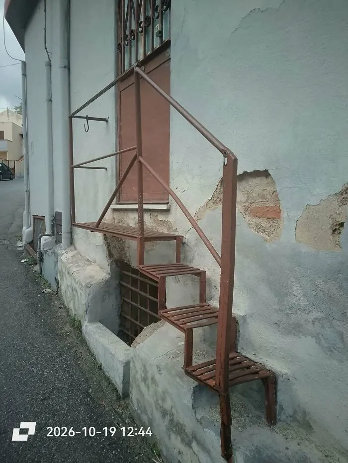 Narrow metal stairs leading to a door on an outdoor wall, showing a construction fail.