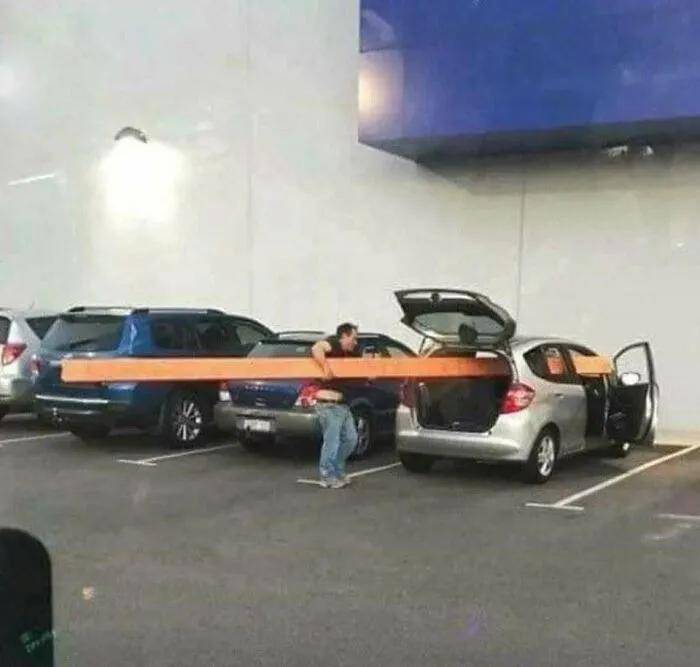 Man attempting to fit an extremely long wooden beam inside a small car, creating a funny construction fail.