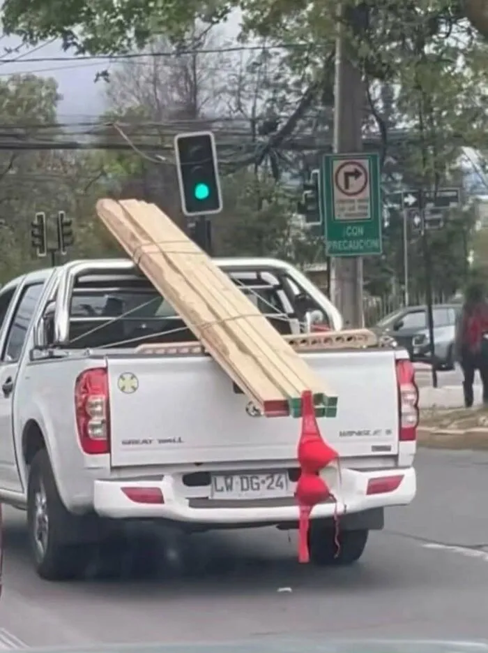 White pickup truck carrying long wooden planks marked with a red bra as a construction fail safety flag.