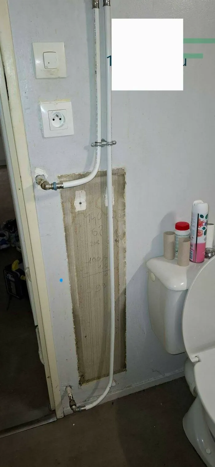 Exposed white plumbing pipe awkwardly running down a bathroom wall next to a toilet, showing a construction fail.