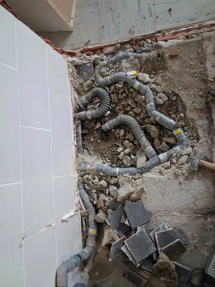 Chaotic mess of exposed grey plumbing pipes laid improperly in a broken floor tile area, highlighting construction fails.