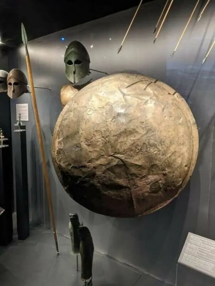 Ancient Greek helmets, spear, and shield displayed in a museum