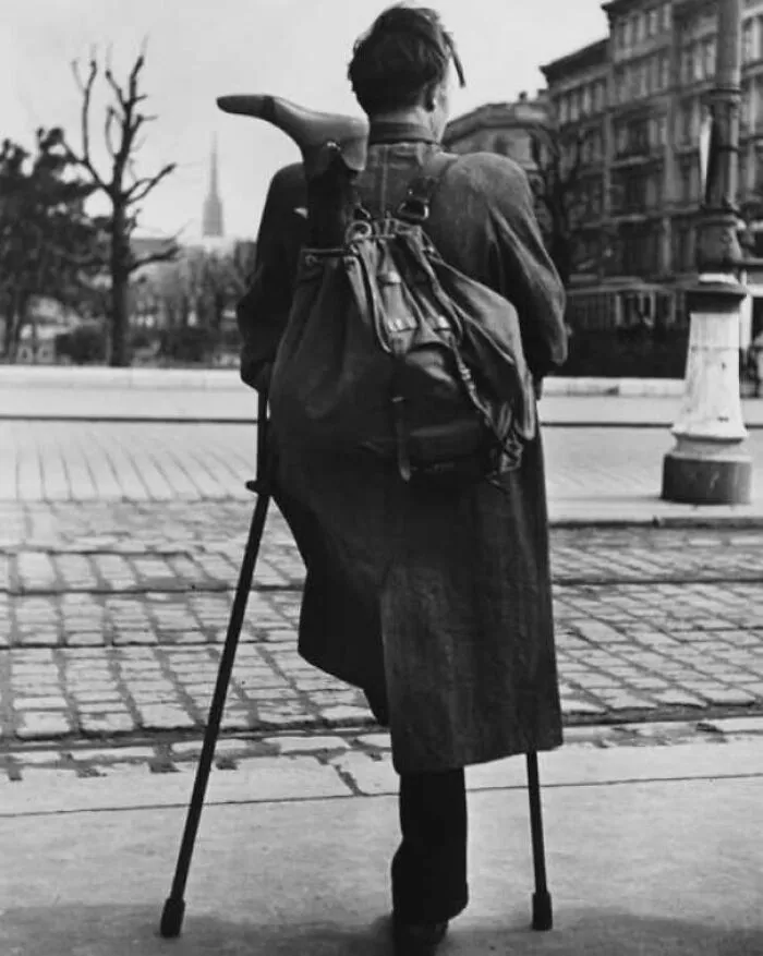 Black and white photo of a person walking on crutches with prosthetic leg in historic urban setting