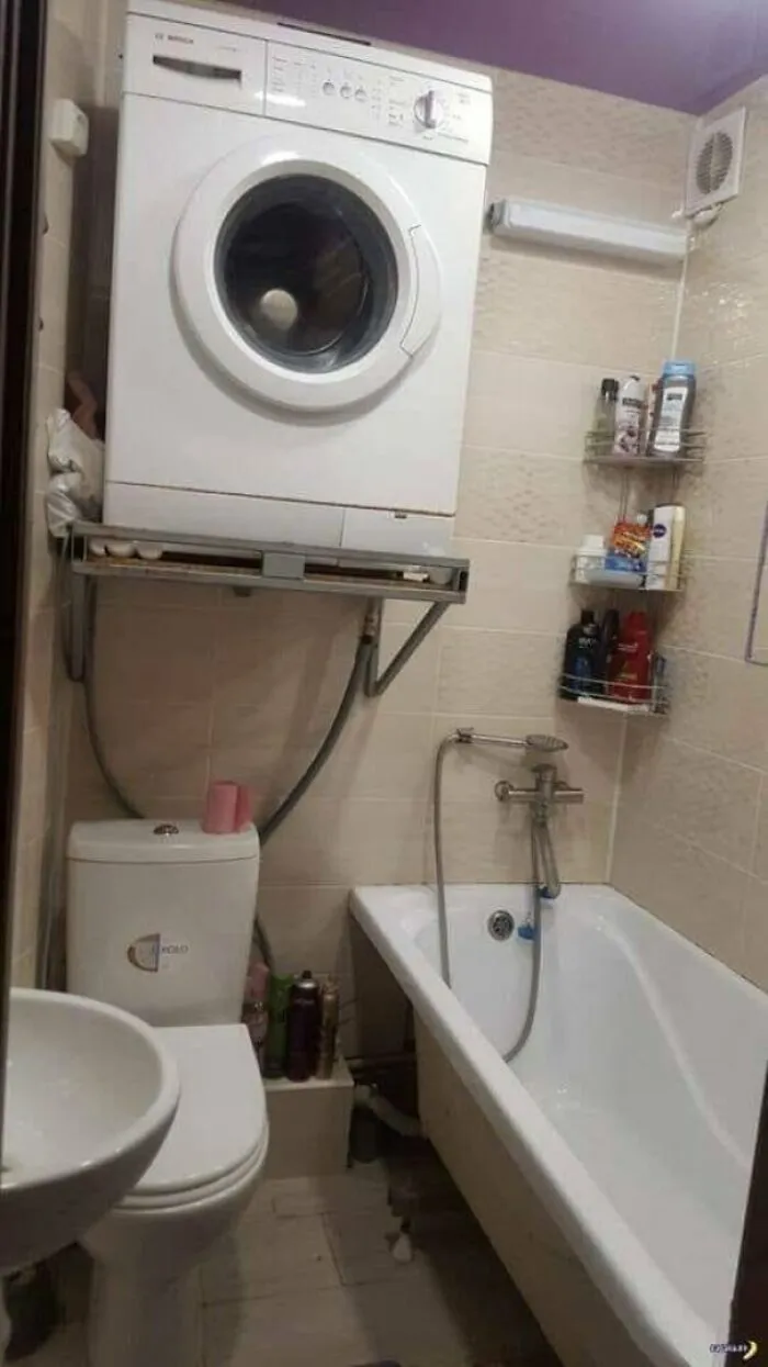A washing machine mounted precariously on a wall shelf above a toilet in a small bathroom