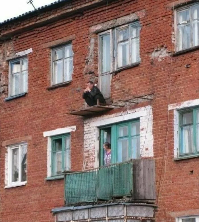 Man squatting dangerously on a tiny ledge outside a window showing wild Slavic engineering.