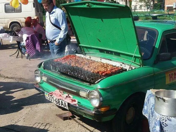 Slavic engineering turns a green car hood into a functional barbecue grill cooking meat at a gathering.