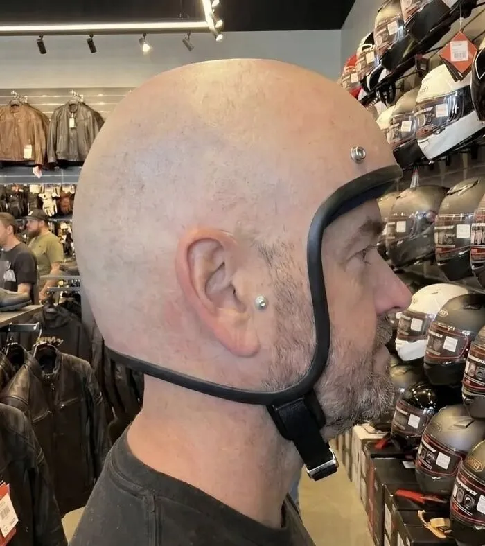 Hilarious pics of a man wearing a realistic bald head motorcycle helmet in a shop