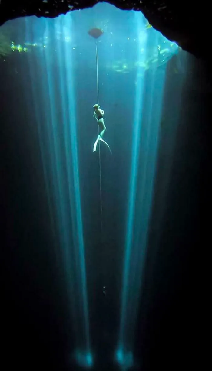 Freediver descending in deep-water cave with sunlight beams piercing through water