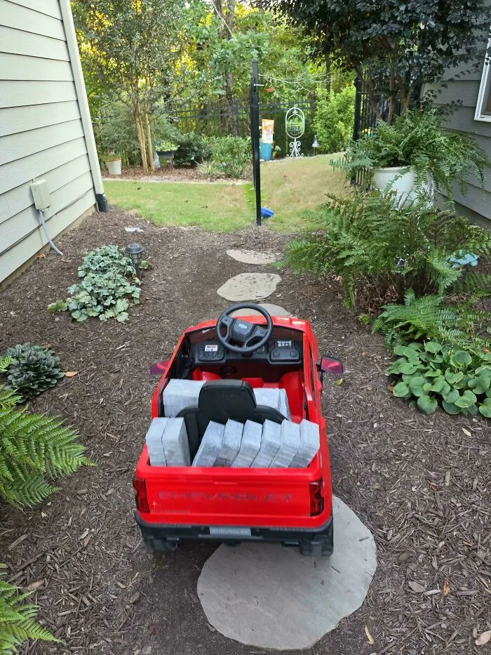 Red toy ride-on truck loaded with pavers in a garden path created by hardworking dads.