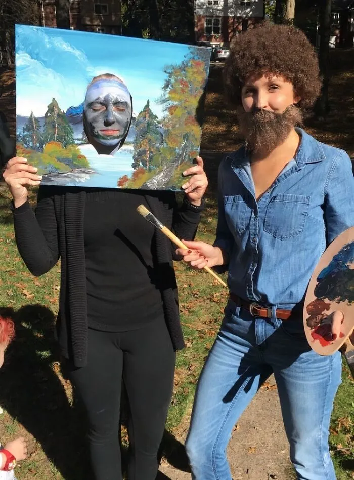 Bob Ross painter and canvas painting as funny Halloween couples costumes posing outside.