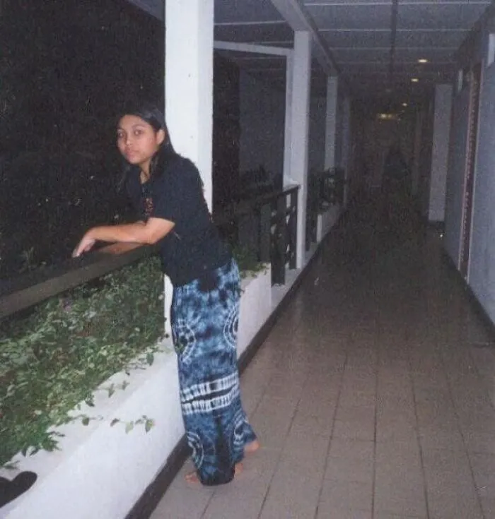 A woman posing on an outdoor walkway with a shadowy figure lurking down the hallway like a horror movie.