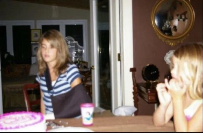 A girl with her arm in a sling sitting at a table with an eerie, shadowy face reflection in a mirror.