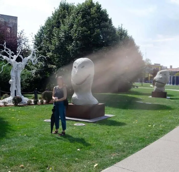 Woman standing in a park next to strange stone head sculptures, giving off horror movie vibes.