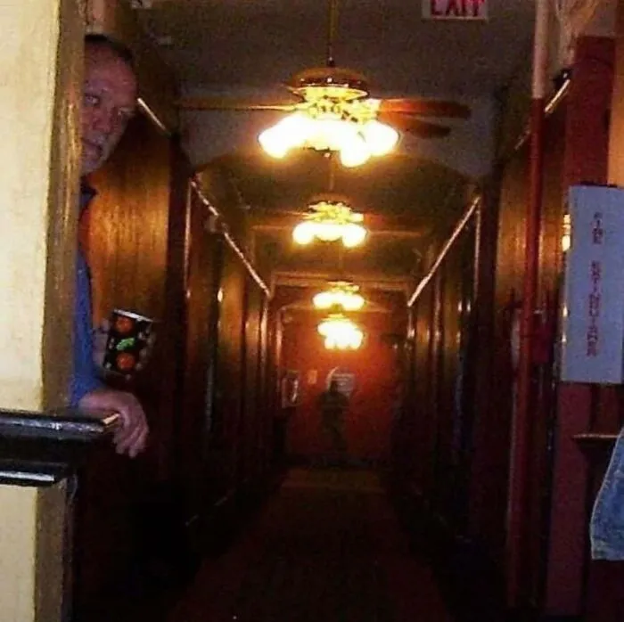 A man peering around a hallway corner with a shadowy figure behind him in a horror movie setting.
