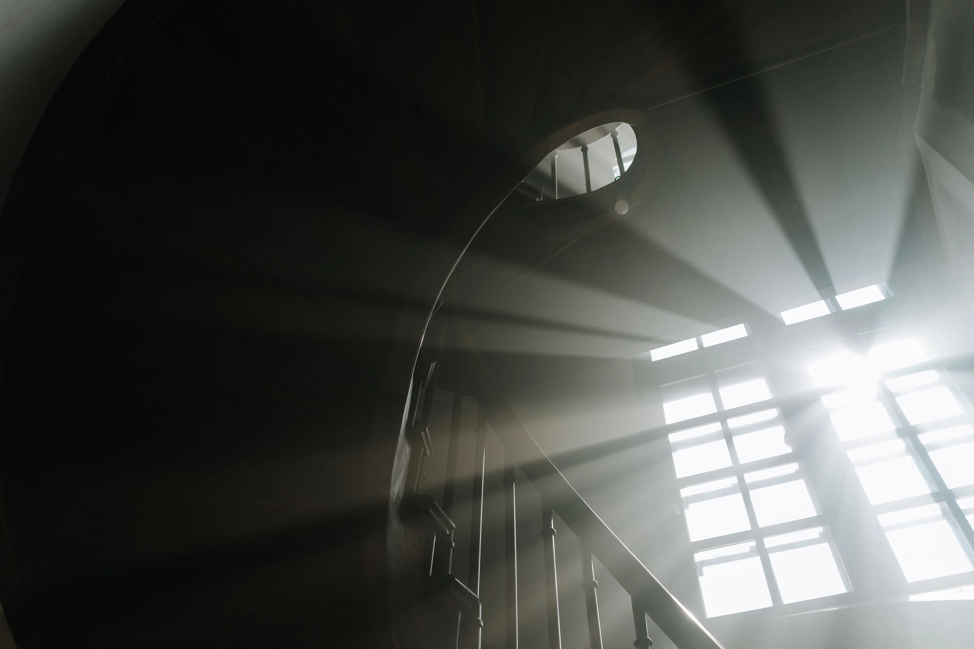 Bright sunlight streams through a window onto a dark wooden spiral staircase.