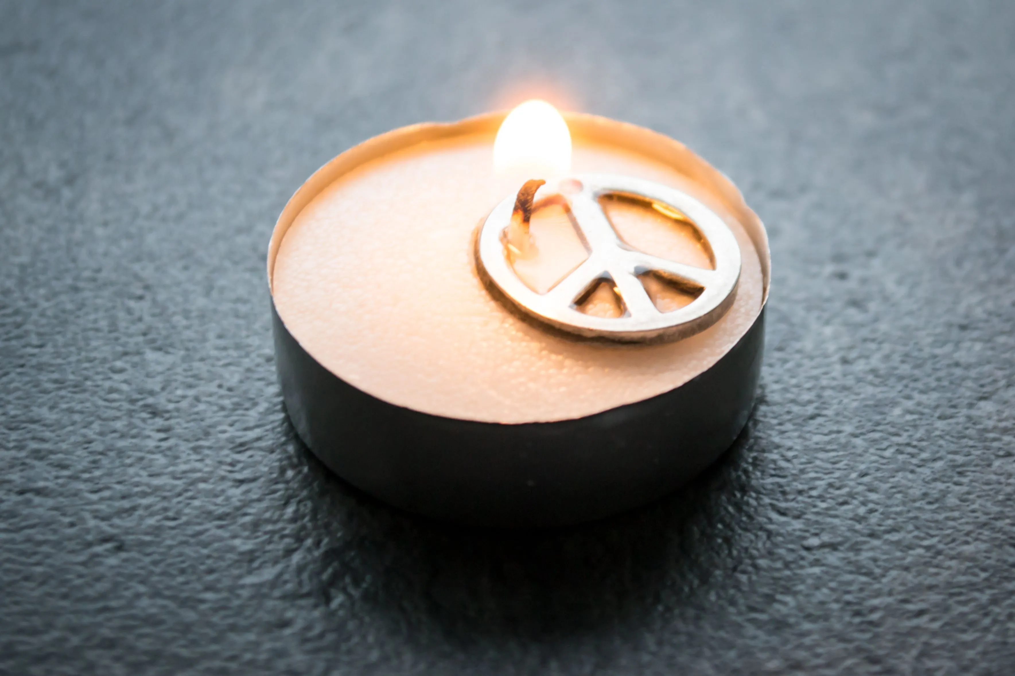 A small lighted tea candle with a metal peace symbol charm resting on top.
