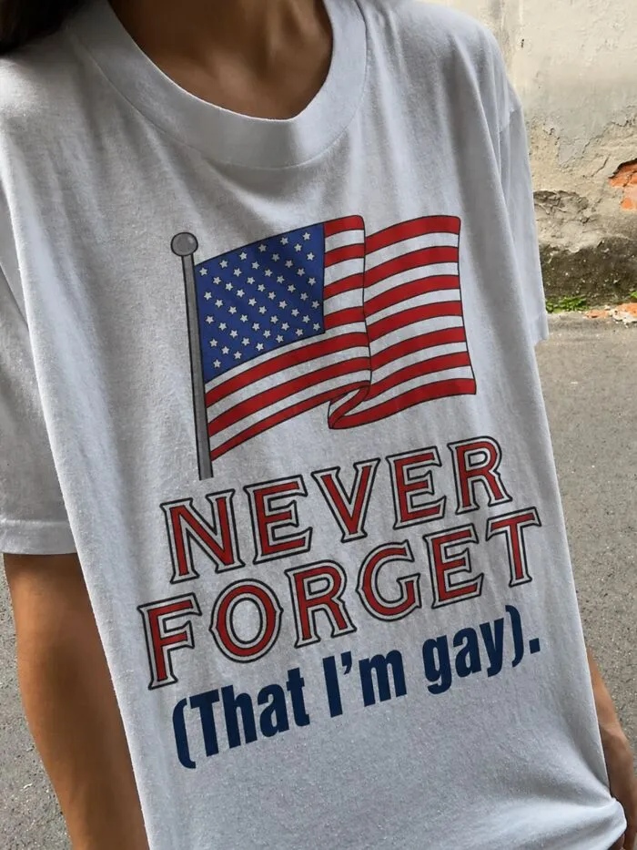 A person wearing a white t-shirt with an American flag graphic and the words NEVER FORGET (That I'm gay).