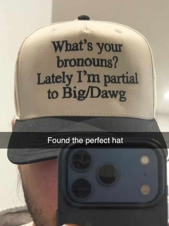 A person wearing a beige and black trucker hat printed with humorous text about bronouns Big and Dawg.