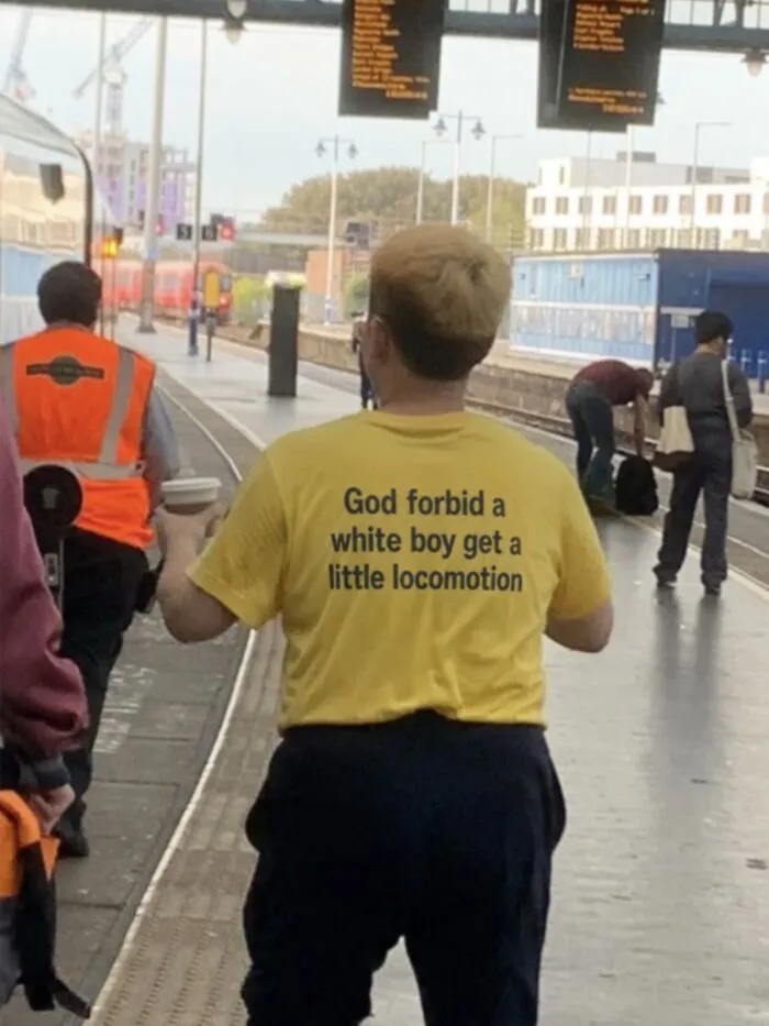 A yellow hilarious t-shirt worn at a train station reading God forbid a white boy get a little locomotion.