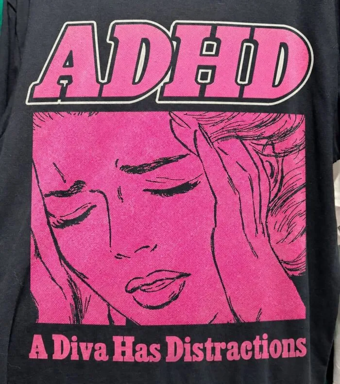 A black t-shirt featuring ADHD and comic artwork of a frustrated woman reading A Diva Has Distractions.