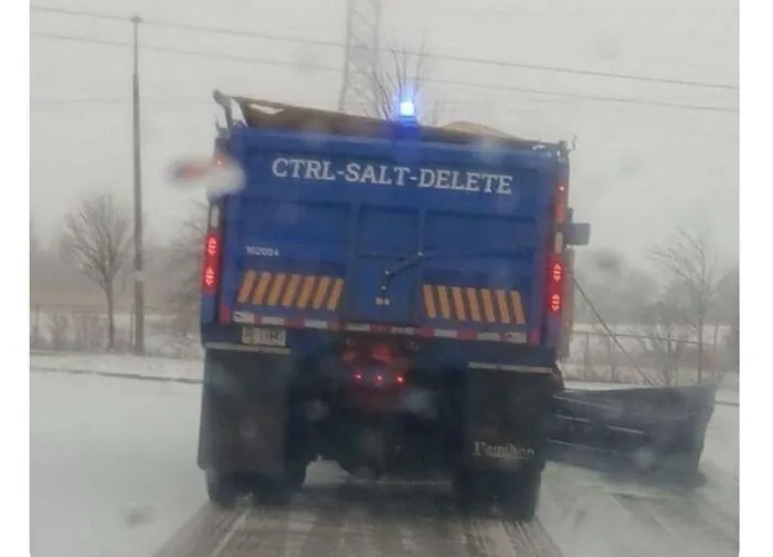 Blue salt truck on a snowy road with a funny joke written on the back reading CTRL-SALT-DELETE