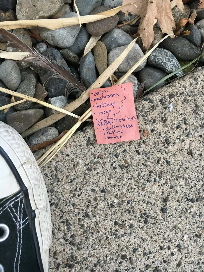 Small pink found paper note with a handwritten grocery list sitting on gravel outdoor ground.