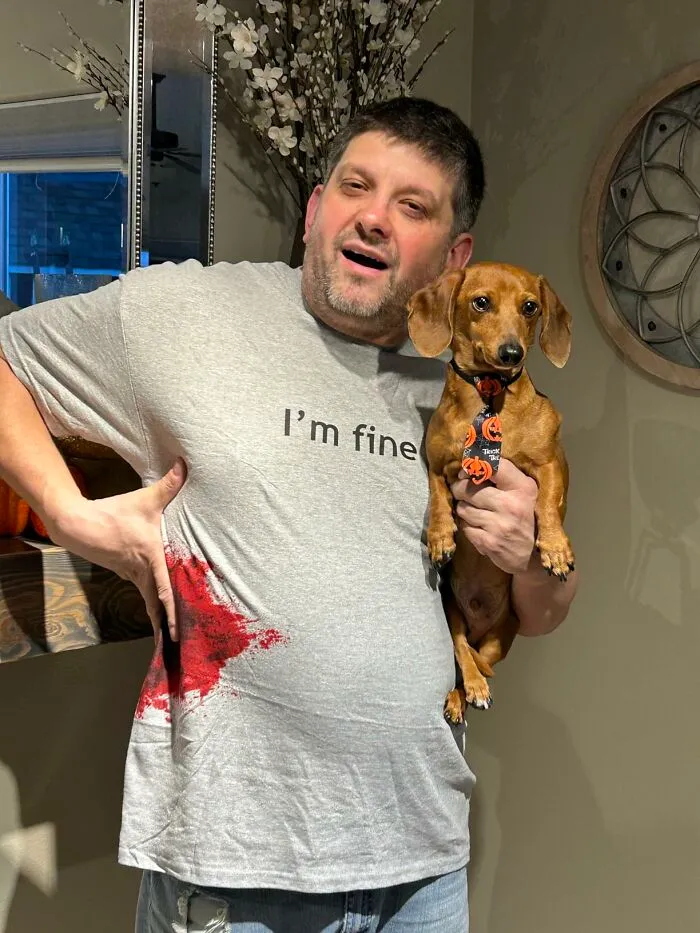 A man wearing a humorous bloody I'm fine shirt holding a dachshund for a low-effort Halloween costume.