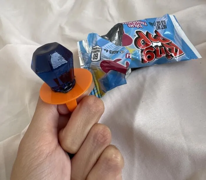 A blue raspberry Ring Pop wearable candy ring held in a hand next to its wrapper for Best Halloween Candies ranking.