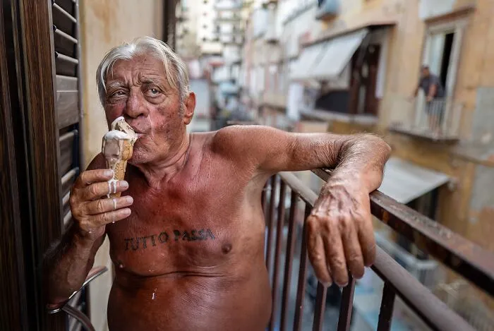 An expressive portrait photo of a shirtless older man on a balcony eating ice cream