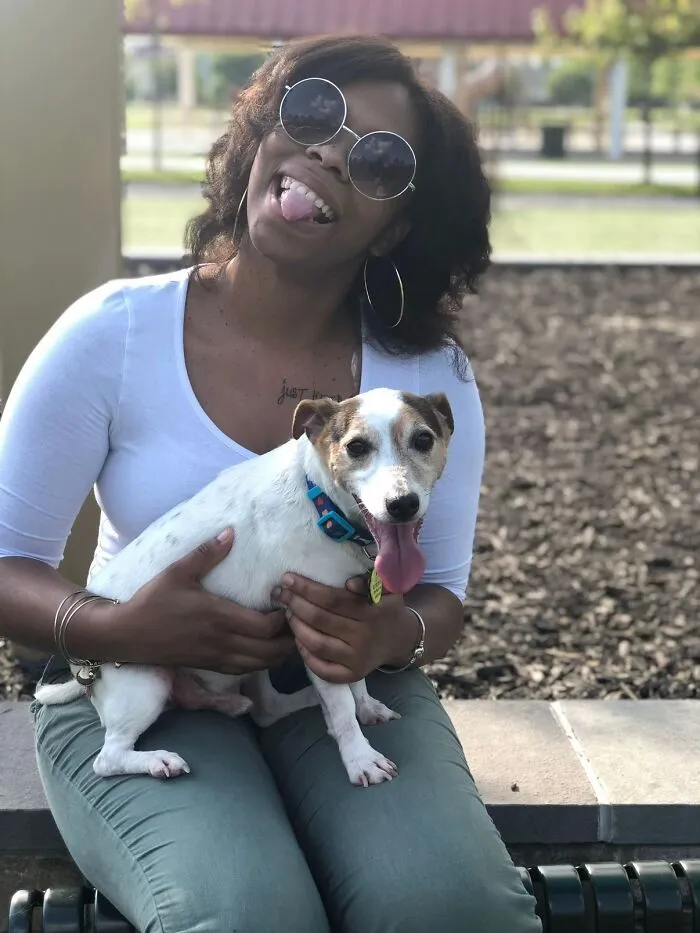 Woman in sunglasses smiles while holding her small dog outside, part of a viral TikTok trend sharing pet stories.